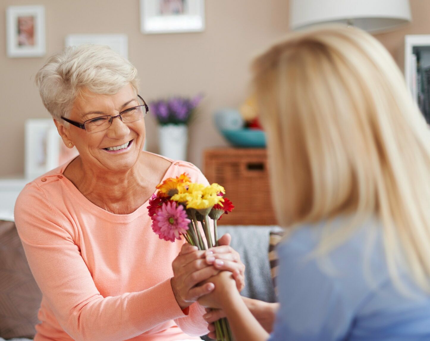 A lady hands a bouquet of flowers to an elderly patient sitting on a sofa